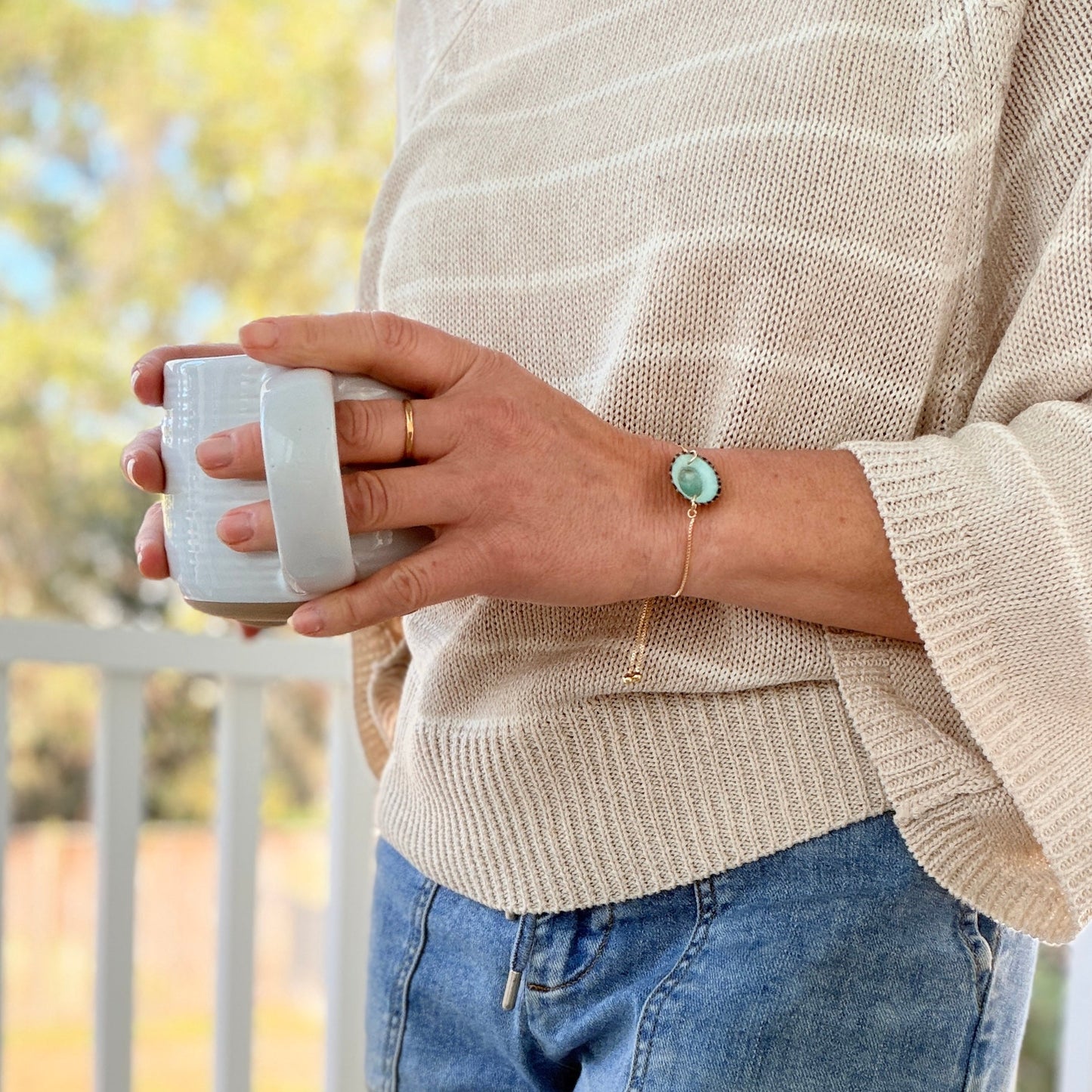 Person wearing a beige sweater and blue jeans holding a white mug outdoors.