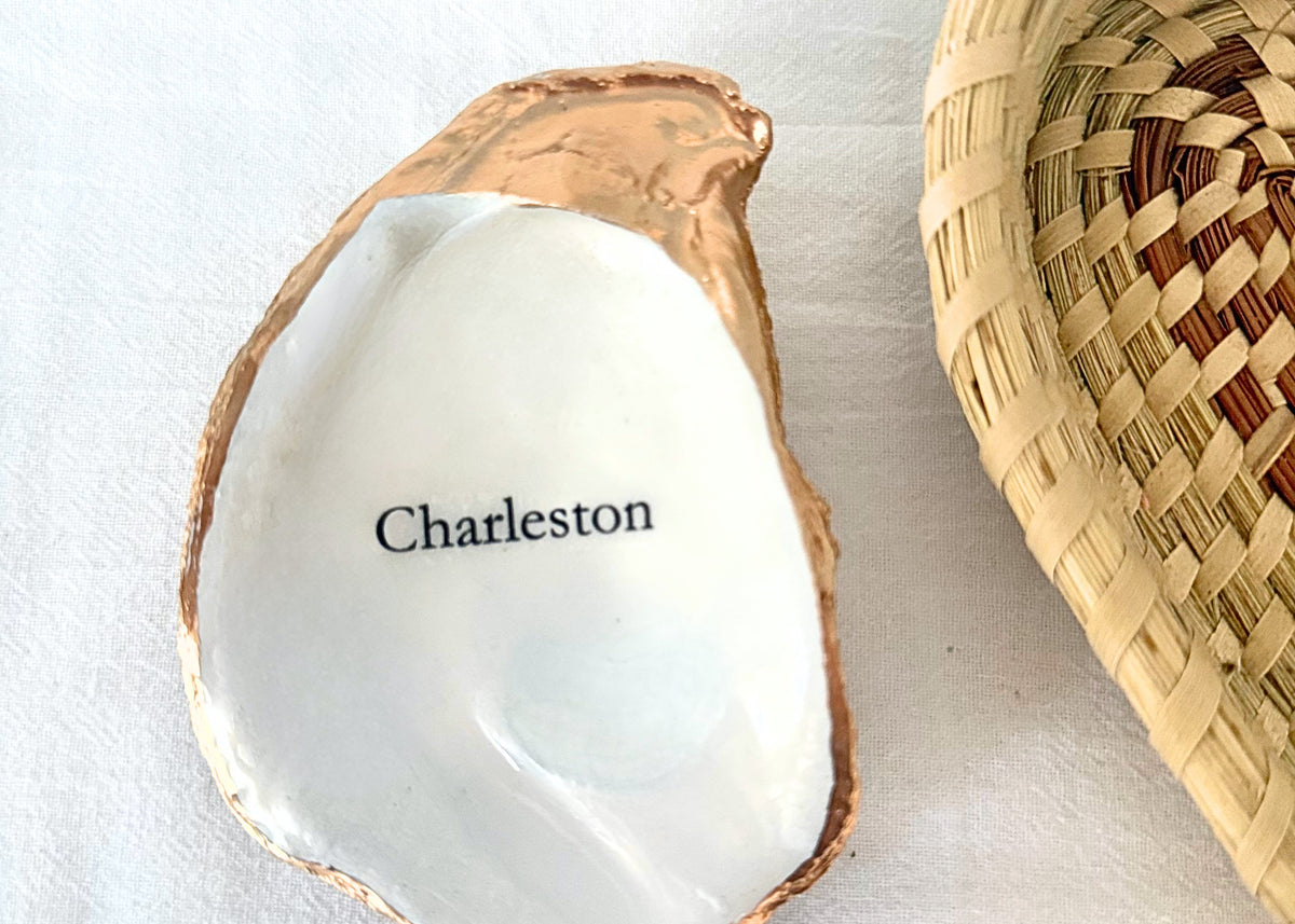 A white ceramic oyster shell dish with a metallic copper-gold rim and the word 'Charleston' printed in black, styled on a white fabric surface next to a woven basket.