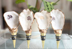 Four oyster shell wine decanters with gold and black metal collars and gold metal stoppers, displayed on a clear acrylic stand with a green plant in the background.